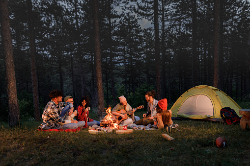 Large group of happy friends having fun while listening to their friend playing a guitar on camping by the bonfire. Copy space.