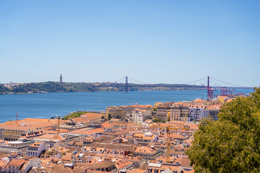 Aerial View of Lisbon, Portugal: A High-Angle, Daytime Capture of the Cityscape, Featuring the Iconic 25 de Abril Bridge Spanning the Tagus River, with a Clear, Blue Sky