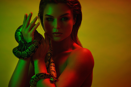Stunning woman in a mix of red and green light, holding two pythons in her hands.