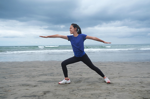 Close-up shot of young Indonesian woman practicing warrior pose at an overcast empty beach