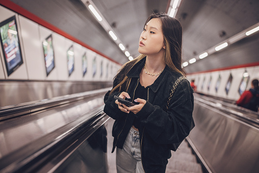 Young Asian woman using mobile phone while riding on escalator in subway station in Vienna.