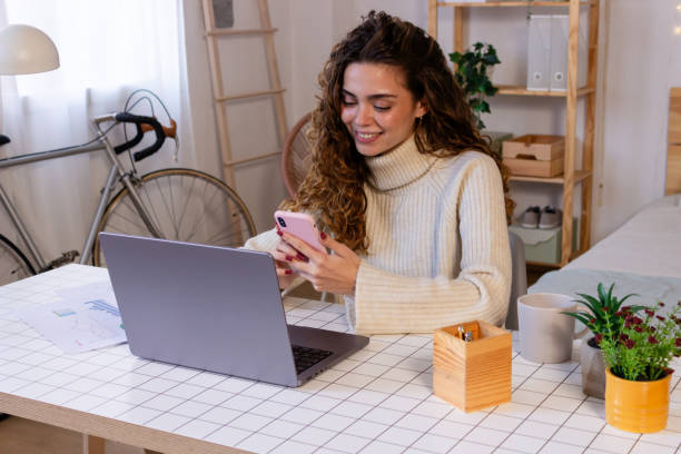 istock Smiling young woman using mobile phone while sitting at table with laptop at home 1851851069