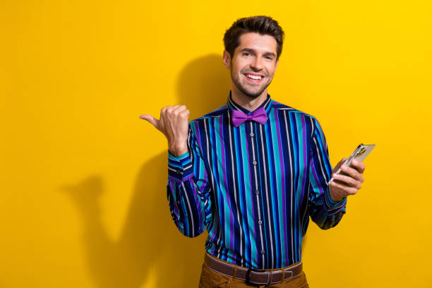 istock Portrait of toothy beaming guy with bow tie hold smartphone directing at eshop empty space isolated on vibrant yellow color background 1851917890