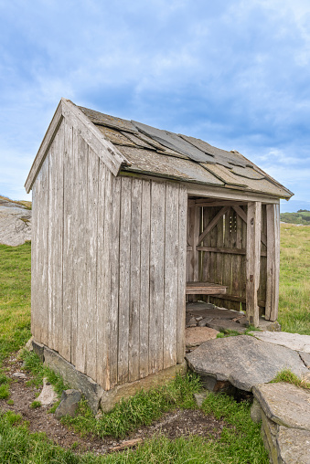 An old traditional rural area worn and weathered bus waiting shed in the countryside of Western Norway in the evening of a summer day. The image was captured with a 24mm wide angle lens with the photographer standing on the country road by the shed. In the background is a landscape of green slopes where sheep are usually grazing.