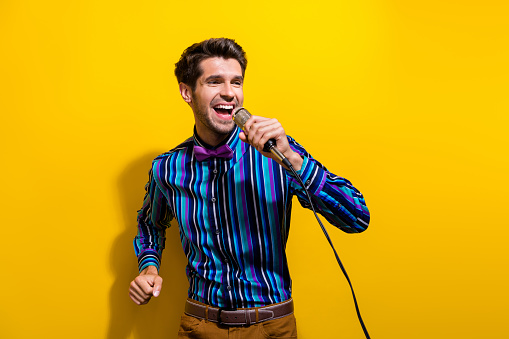 Portrait of cheerful guy dressed stylish shirt singing pop song in microphone at retro party isolated on bright yellow color background.
