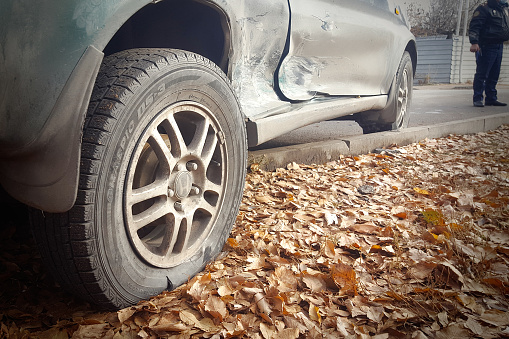 A dented car with a damaged door and a torn wheel stands on the side of the road after an accident with a police officer in the background. Almaty, Kazakhstan - October 11, 2021