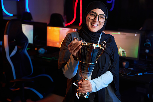 Waist up portrait of Muslim young woman holding golden cup and smiling at camera celebrating victory in eSports championship, copy space