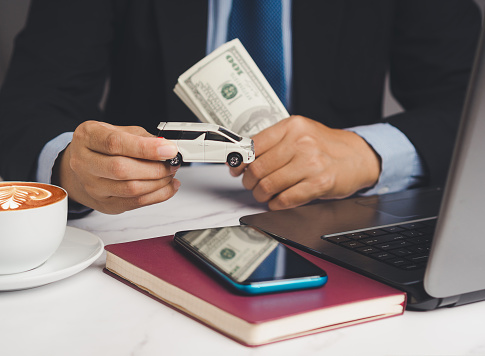 Car loan or Title loan. US dollar bills in a hand businessman while sitting at the table. Miniature a white car model, a calculator, and a laptop on a table. Car finance and insurance concept