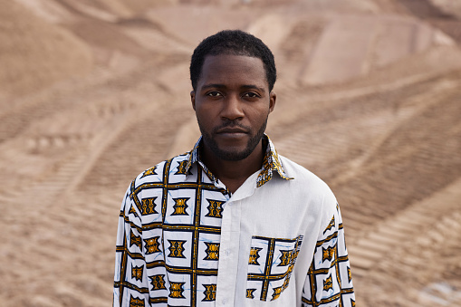 Waist up portrait of handsome Black man wearing white shirt with traditional ornaments in desert landscape and looking at camera