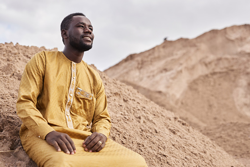 Low angle portrait of smiling Black man wearing traditional kaftan looking at view in desert on sand dune, copy space