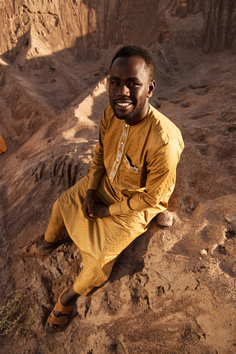High angle portrait of smiling Black man sitting on sand dune and looking up at camera in sunlight
