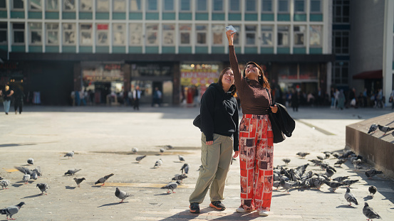 Two multi-ethnic female tourist friends are using a mobile smart phone to take selfies in a city.