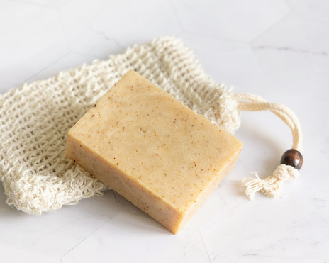 Beige handmade soap bar on sisal soap saver bag on white marble table close up, copy space. Natural herbal products for face and body care