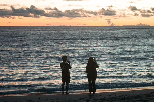 Rear view silhouette photo of young couples watching and taking photos of sunset, on the beach. with Orange sunet sky as background