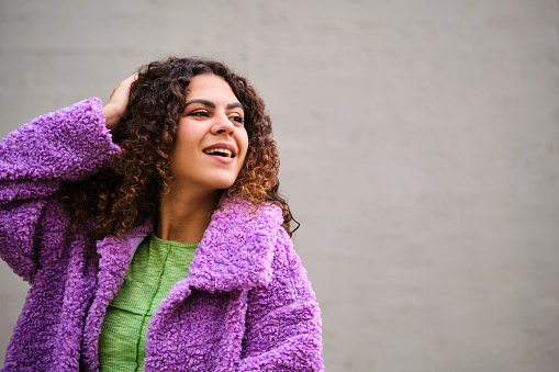Young Spanish woman with curly hair and purple coat posing looking at a side over gray wall.