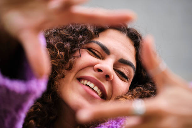 Young Spanish woman with curly hair smile and make a frame with hands. Young Spanish woman with curly hair and purple coat smiling and making a frame with hands over gray wall. selfie madrid photos stock pictures, royalty-free photos & images