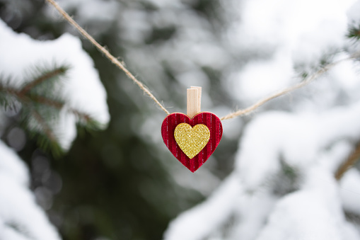 Red Christmas ornament in shape of heart hanging on jute twine string on snow-covered fir tree background outdoors. Concept of love, family, support