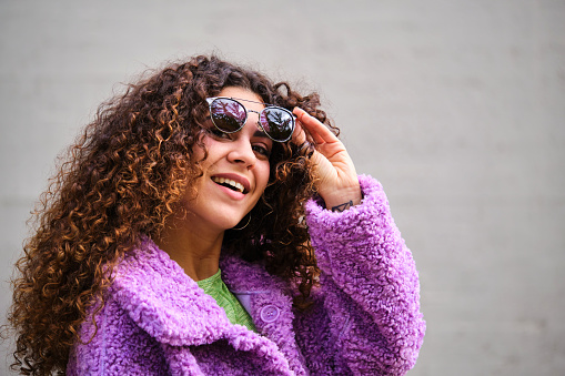 Young Spanish woman with curly hair, purple coat and sunglasses smiling and looking at camera over gray wall.