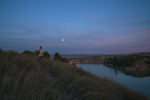 Latin photographer with a camera takes photos in a lagoon and the moon at dusk