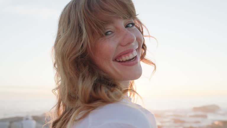 Portrait, flare and a woman laughing on the beach at sunset for travel, freedom or inspiration by the ocean. Face, smile and funny with a young redhead person on the coast in summer for vacation