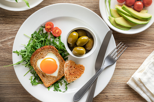 Heart shaped egg in a toast with olives and vegetables top view on plate, romantic healthy breakfast