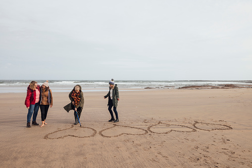 A wide angle view of a group of women enjoying a walk along the coast in Bamburgh in the North East of England. One of the women who has recently overcame breast cancer is drawing a heart in the sand as she stands with her friends. She is drawing a heart for each one of them.