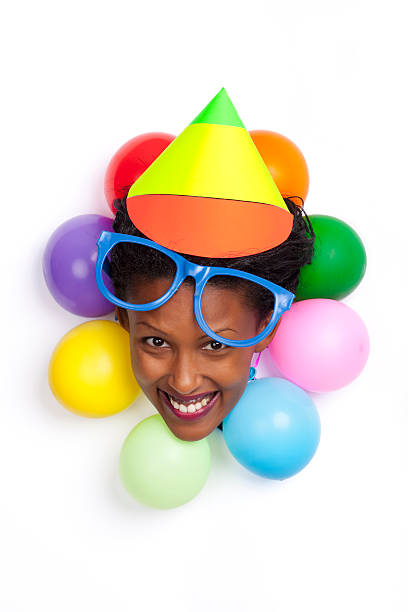 Portrait of funny clown woman face with colored balloons around his neck, huge blue  eyeglasses and colored cone hat on his head. Head peering out hole in paper.