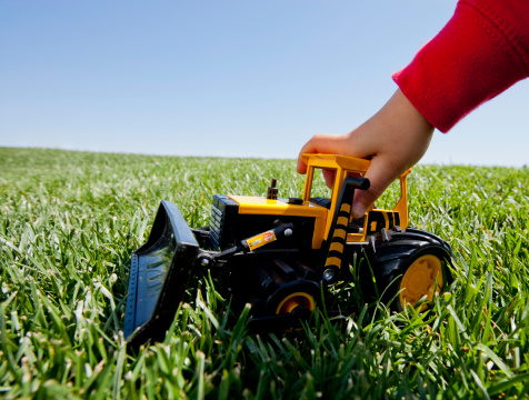 A boy with a red sweatshirt is playing with a toy front loader bulldozer on the grass during the day.