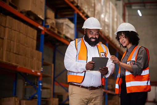 Two smiling engineers illuminated by the glow from a digital tablet screen while tracking orders in a large shipping and distribution warehouse