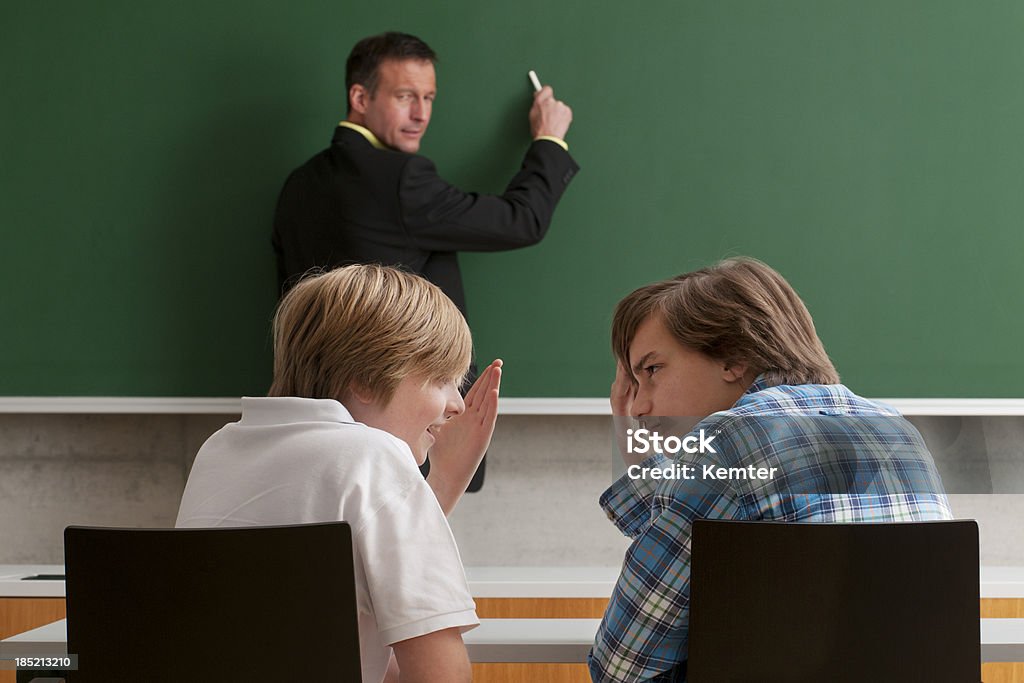 pupils whispering during lesson students whispering while teacher is writing on blackboard - focus on students 40-49 Years Stock Photo
