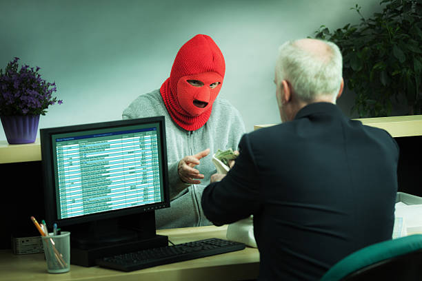 A violent criminal bank robber wearing a red ski mask and gray jacket holding a concealed weapon in his pocket is in the process of a bank robbery in a retail bank counter terminal. Frightened bank teller is in a fearful confusion offering cash to the robber. Photographed in a horizontal format in low key lighting.