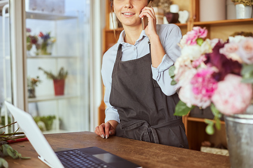 Portrait of florist in apron working at her own flower shop, using laptop and calling on the smart phone. She is leaning on wooden counter. She is arranging logistics, and delivery, taking orders