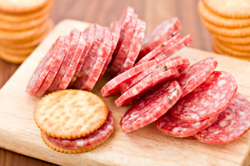 A high angle close up shot of a wooden cutting board with sliced salami and a slice of salami between two crackers. Two stacks of crackers sit on the foreground