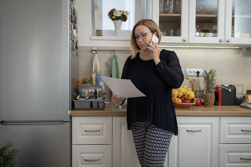 Shot of an attractive adult woman reading paperwork and talking on a phone while working from home