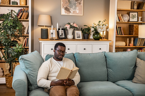 Handsome smiling black man reading a book while sitting on the couch in the living room, enjoying a rest day