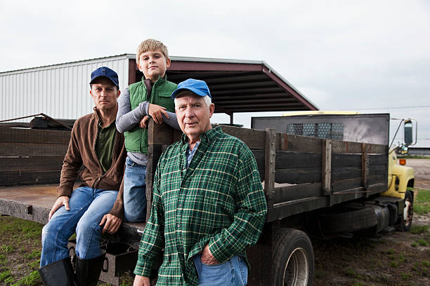 Multi-generation family working on the farm Three generations of men at the back of a truck on the family farm.  The grandfather, a senior man in his 60s, is standing with his hand in his pocket, looking at the camera.  His adult son and young grandson are behind him on the truck.  They are wearing jeans, work boots and jackets or flannel shirt.  Everything is brown or green, earthy colors. group of people men mature adult serious stock pictures, royalty-free photos & images