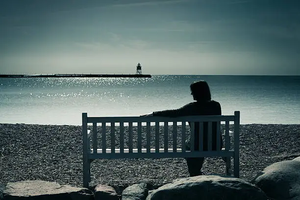 Woman in silhouette, sitting on a bench in a moody landscape at water's edge. The lone woman may be widowed, divorced, or lonely, sitting in solitude, contemplating her grief, sadness, or depression. The lighthouse in the distance is a symbol of finding hope or direction in a sea of troubles.