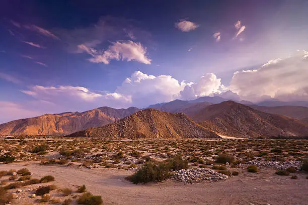 Flat desert quickly gives way to steep dry hills in the dusty Southern California desert, set against a dramatic sky.