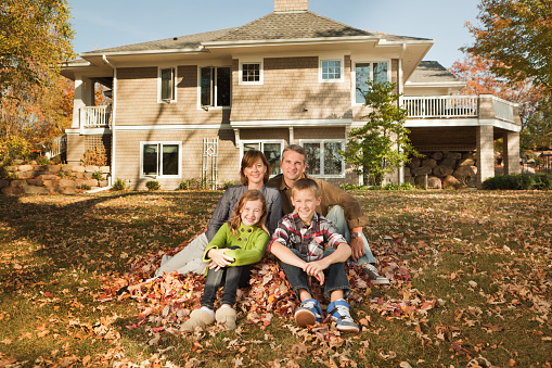 Family of four sitting together outdoors in a raked autumn leaf pile, in front of their residential building home. The large suburban house and lawn provide an idyllic lifestyle for cheerful, happy parents with two children, boy and girl. A casual, real people portrait of family togetherness, love and affection.