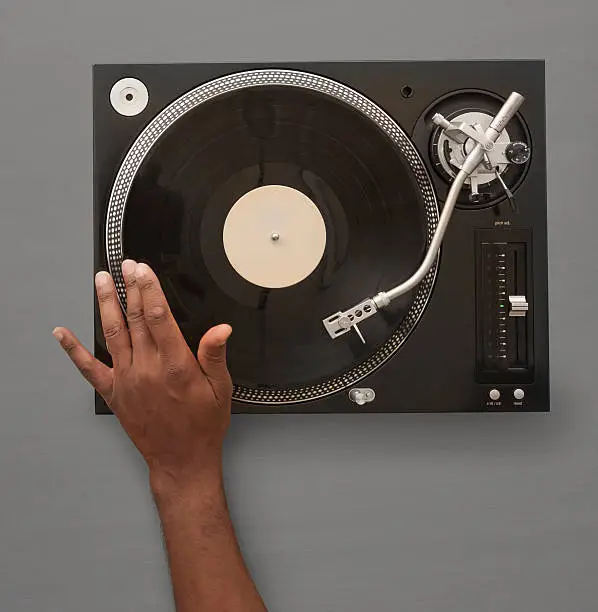 An overhead photo of a male African American hand spinning or playing a record on an record player / Turn Table.  Logos have been removed from player.