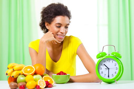 Mixed race woman looking at clock, sitting by the table full of fresh fruits.