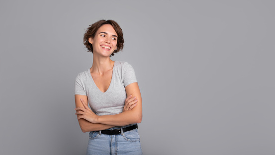 Close up photo of beautiful smiling happy young girl with short hair and crossed arms, in casual wear, looking away. Studio shot of cute woman isolated against blank gray wall.