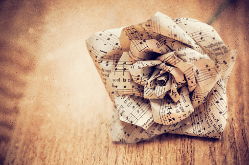 A handmade paper craft shaped to look like a rose laying on a wooden floor surface.  Made from old hymn sheets.  Overlayed with grunge paper for a vintage feel.