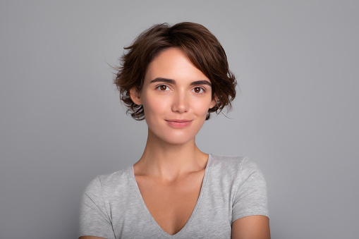 Close up portrait of pretty smiling happy woman with short hair, in casual wear, looking calmly into camera. Studio shot of gorgeous girl isolated against blank gray wall.