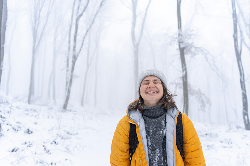 Happy young caucasian woman in yellow jacket enjoying snowfall in winter forest, exposing her face to snowflakes