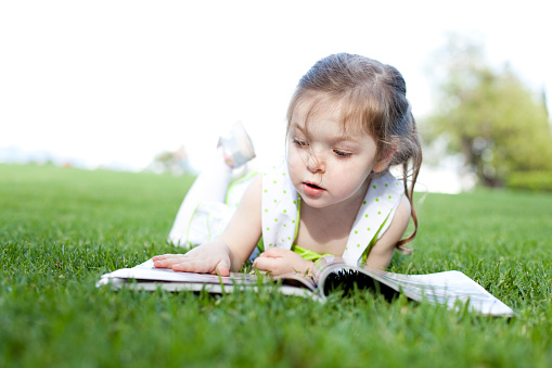 Little caucasian girl lying on the grass at park and reading magazine