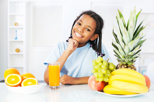 Little cheerful girl with orange juice and fruit leaning on the table.