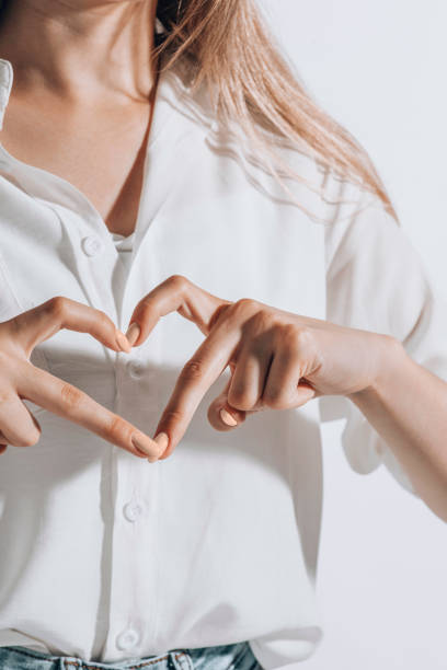 romantic young woman in shirt making a heart gesture with her fingers in front of her chest showing her love and affection stock photo