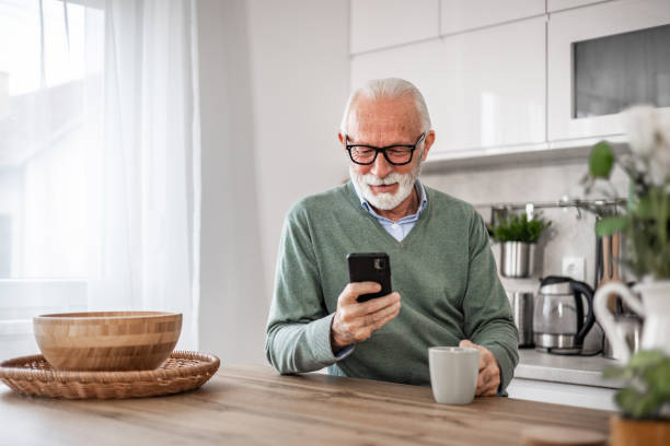 istock Old man, pensioner, sitting in kitchen with happy smile drinking hot coffee or tea and using mobile phone, morning routine, carefree retirement days 1852316937