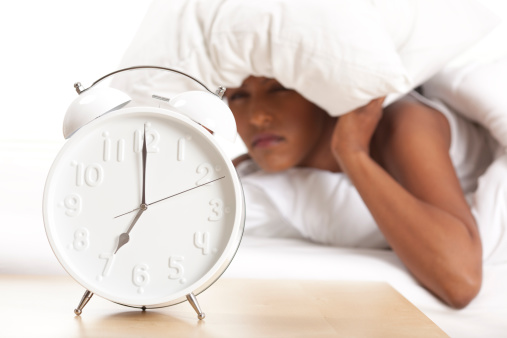 Tired woman covers her head with a pillow after the alarm clock rings.